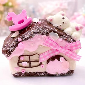 May include: A miniature house with a chocolate-covered roof, decorated with a pink rocking horse and a white bear. The house has pink details, a gingham bow, and a window. The design is whimsical and charming, with a sweet aesthetic.