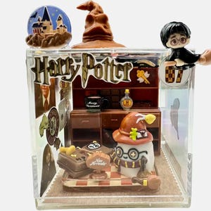 May include: A clear, square display case featuring a miniature Harry Potter scene. The case includes a Sorting Hat, Hogwarts castle, and a figure of Harry Potter on a broom. The words "Harry Potter" are displayed on the front.