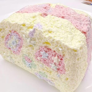 May include: A colorful, frosted cake roll with a pastel rainbow design. The cake features layers of pink, yellow, and white frosting, with decorative star-shaped sprinkles. The cake is a dessert item.