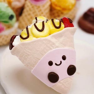 May include: A cream-coloured, cone-shaped dessert with yellow and red toppings, drizzled with chocolate. A pink, smiling face with brown eyes and a mouth is attached to the cone. Other similar desserts are in the background.