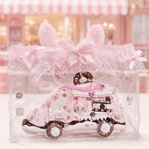 May include: A clear plastic bag with a pink lace trim and two fuzzy pink ears on top. Inside is a pink food truck figurine featuring a My Melody character, chocolate-coloured accents, and various small decorative stickers of sweets and stars.