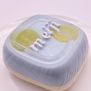 May include: A square-shaped pastry with a light blue top and a white base. The top features the word "meiji" in white script and two yellow-green shapes. The pastry is presented on a white plate.