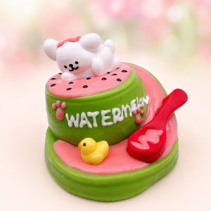 Peut inclure: Une figurine miniature sur le thème de la pastèque, verte et rose, avec un ours blanc en dessin animé allongé sur le dessus. La base verte porte le mot WATERMELON en lettres blanches, un petit canard jaune et une cuillère rouge.