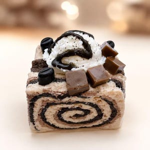 May include: A square-shaped dessert block featuring a swirled pattern of light tan and dark brown layers. The top is decorated with a dollop of white whipped cream, dark chocolate drizzle, three dark coffee beans, and three brown caramel cubes.