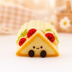 May include: A charming, yellow sandwich-shaped ornament with a smiling face and black wheels. The sandwich has red tomato slices and green lettuce, with a golden-brown crust. A cute, food-themed decorative item.