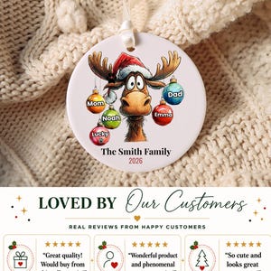 K&ouml;nnte beinhalten: Ein runder, wei&szlig;er Keramik-Weihnachtsanh&auml;nger mit einem Cartoon-Elch, der eine Weihnachtsm&uuml;tze tr&auml;gt. Der Elch ist von bunten Kugeln umgeben, die mit Mom, Noah, Lucky, Emma und Dad beschriftet sind. Der Text lautet The Smith Family und 2026.