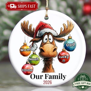 K&ouml;nnte beinhalten: Ein rundes, wei&szlig;es Keramik-Weihnachtsornament mit einem Cartoon-Elch, der eine Weihnachtsmannm&uuml;tze tr&auml;gt. Der Elch hat f&uuml;nf bunte, h&auml;ngende Kugeln mit den Aufschriften Mom, Noah, Emma, Lucky und Dad. Unten steht der Text Our Family 2026.