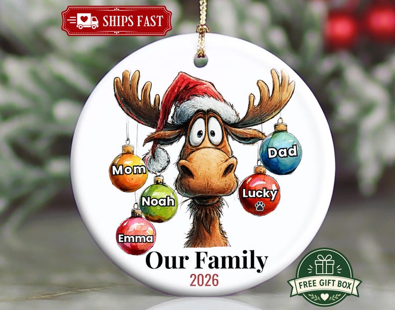 K&ouml;nnte beinhalten: Ein rundes, wei&szlig;es Keramik-Weihnachtsornament mit einem Cartoon-Elch, der eine Weihnachtsmannm&uuml;tze tr&auml;gt. Der Elch hat f&uuml;nf bunte, h&auml;ngende Kugeln mit den Aufschriften Mom, Noah, Emma, Lucky und Dad. Unten steht der Text Our Family 2026.