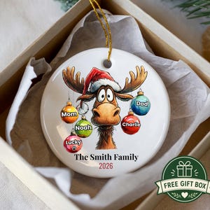 K&ouml;nnte beinhalten: Ein rundes, wei&szlig;es Keramikornament mit einem Cartoon-Elch, der eine rote Weihnachtsm&uuml;tze tr&auml;gt. Der Elch ist von bunten Weihnachtskugeln umgeben, die mit den Namen Mom, Noah, Lucky, Charlie und Dad beschriftet sind. Der Text lautet The Smith Family 2026.