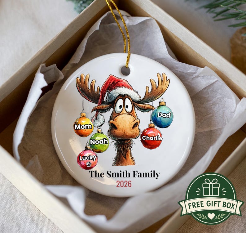 K&ouml;nnte beinhalten: Ein rundes, wei&szlig;es Keramikornament mit einem Cartoon-Elch, der eine rote Weihnachtsm&uuml;tze tr&auml;gt. Der Elch ist von bunten Weihnachtskugeln umgeben, die mit den Namen Mom, Noah, Lucky, Charlie und Dad beschriftet sind. Der Text lautet The Smith Family 2026.