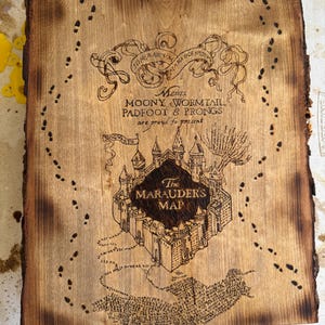 May include: A wooden plaque with a pyrography design of The Marauder's Map from the Harry Potter series. The map features the Hogwarts castle, footprints, and text including "Moony, Wormtail, Padfoot & Prongs". The edges of the plaque are dark brown.
