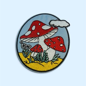 Op de afbeelding: Een ronde geborduurde patch met drie rood-witte stippenpaddenstoelen op een gele ondergrond. De achtergrond bevat een lichtblauwe lucht met een witte wolk, kleine groene varens en witte bloemen, alles omrand door een zwarte rand.