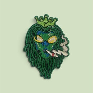 May include: An embroidered patch featuring a green-skinned figure with long green dreadlocks wearing a green crown. The figure has yellow eyes, a blue nose, and is smoking a white cigarette with grey smoke swirling to the right side.