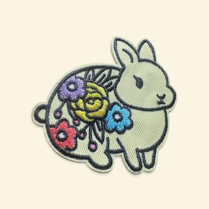 Op de afbeelding: Een geborduurde patch in de vorm van een lichtgroen konijn. Het konijn heeft een decoratief bloemenpatroon op de rug met geborduurde bloemen in tinten geel, rood, blauw en paars, allemaal omlijnd met een donkerzwarte draad.
