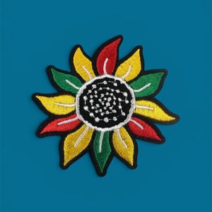 Op de afbeelding: Geborduurde zonnebloemplakker met een zwarte rand. De bloemblaadjes zijn geel, rood en groen en omringen een zwart midden met witte stiksels. De patch is geplaatst tegen een effen blauwgroene achtergrond.