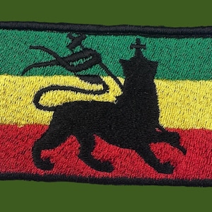 Op de afbeelding: Geborduurde patch met een zwart leeuwsilhouet met een kroon, tegen een achtergrond van groene, gele en rode strepen. De patch heeft een zwarte rand en een getextureerde uitstraling.