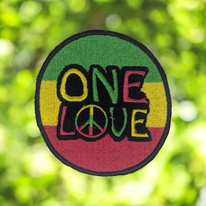 Può includere: Toppa ricamata circolare con le parole "ONE LOVE" in nero, verde, giallo e rosso. La toppa include un simbolo di pace. Lo sfondo è verde sfocato, che suggerisce un ambiente naturale.