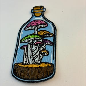 Op de afbeelding: Geborduurde patch met kleurrijke paddenstoelen in een glazen fles. De patch heeft een blauwe achtergrond, een bruine basis en een kurken dop. De paddenstoelen hebben roze, gele, groene en paarse hoeden.