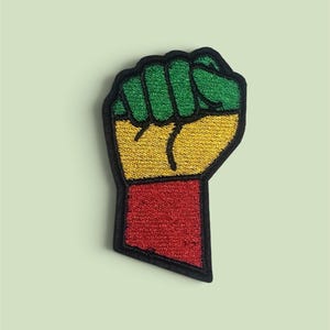 Può includere: Una toppa ricamata a forma di pugno alzato. Il design presenta blocchi di colore orizzontali con verde in alto, giallo al centro e rosso in basso. L'intera forma del pugno è delineata da un bordo nero spesso.