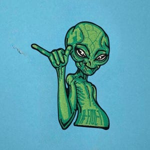 May include: An embroidered patch featuring a green alien with a large head and dark eyes, making a shaka hand gesture. The alien has a textured, light and dark green skin pattern and is outlined in black thread against a solid light blue background.