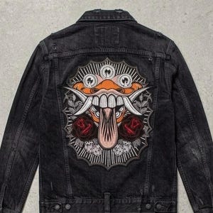 May include: A black denim jacket featuring a large back embroidery of a stylized creature with orange skin, white fangs, and a long tongue. The design includes two red roses, dice, and circular emblems with axe icons, all framed by a white sunburst border.