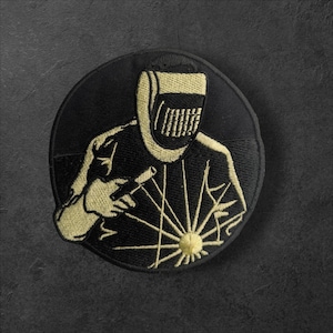 Op de afbeelding: Geborduurde ronde patch met een lasser in een beschermend masker en handschoenen, die een lasbrander vasthoudt. Het ontwerp is in goud en zwart, met een gedetailleerde weergave van lasvonken.