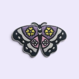 Op de afbeelding: Een geborduurde vlinderpatch met een symmetrisch ontwerp met paarse en roze vleugels, gele bloemaccenten en een zwarte rand met witte stippen. De vlinder heeft een zwart lichaam met witte markeringen en twee dunne voelsprieten aan de bovenkant.