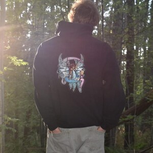 May include: Black hooded sweatshirt with a colourful graphic on the back. The graphic features a winged figure with a crown and other fantasy elements. The person is wearing grey trousers and standing in a forest.