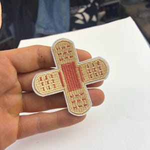 May include: Embroidered patch in the shape of a cross, resembling a bandage. The patch features a tan background with a red center and white border. The design is detailed with small, repeating patterns.