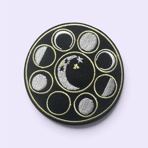 May include: A circular black embroidered patch featuring a central crescent moon with small stars and a surrounding border of eight distinct lunar phases. The entire design is outlined in gold thread, creating a detailed celestial motif.