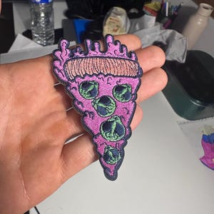 Op de afbeelding: Een driehoekige geborduurde patch in de vorm van een pizzapunt met een roze korst en roze kaas. De pizzapunt is voorzien van drie groene alienhoofden met grote zwarte ogen als topping, allemaal omlijnd met donker garen en een textuur.