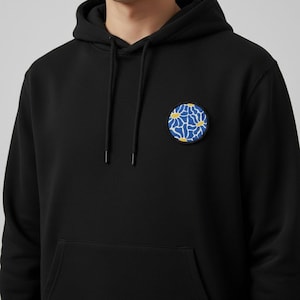 Op de afbeelding: Zwarte hoodie met een ronde patch met een blauw en wit bloemenontwerp met gele accenten. De hoodie heeft een capuchon met trekkoord en een voorzak.