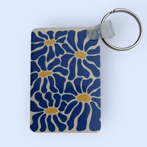 Op de afbeelding: Rechthoekige sleutelhanger met een bloemenpatroon. Het ontwerp toont blauwe bloemvormen met gele centra op een beige achtergrond. Een zilveren sleutelring en een plastic sluiting zijn bevestigd.