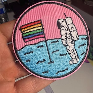 Op de afbeelding: Een ronde geborduurde patch met een witte astronaut die op een getextureerd blauw oppervlak staat tegen een roze achtergrond. Een vlaggenmast met een horizontale regenboogvlag staat in het blauwe oppervlak naast de astronaut.