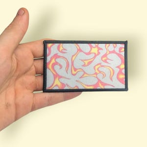 Op de afbeelding: Een rechthoekige patch met een grijze achtergrond en een herhalend vlammenpatroon in tinten geel en roze. De patch heeft een zwarte rand.