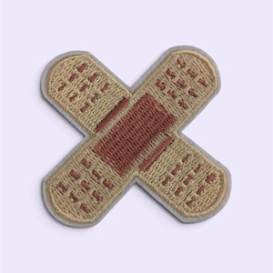 Op de afbeelding: Een geborduurde patch in de vorm van twee gekruiste pleisters. Het ontwerp heeft een beige basis met een donkerbruin vierkant in het midden en kleine bruine rechthoekige accenten aan de uiteinden, omlijnd met een lichtgrijze rand.