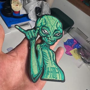 Op de afbeelding: Een geborduurde patch met een groen alien-figuur dat een shaka-handgebaar maakt. De alien heeft grote donkere ogen, een textuur op het hoofd en een torso met de tekst "HIDE" geborduurd in een vet lettertype. De patch heeft een zwarte rand.
