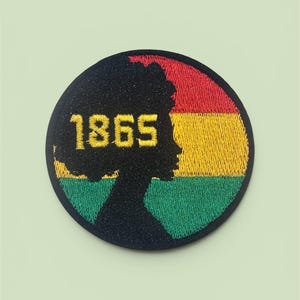 Puede incluir: Un parche bordado circular que presenta la silueta negra de una persona con peinado afro. El fondo consta de franjas horizontales rojas, amarillas y verdes. El número 1865 en amarillo está bordado sobre la silueta.