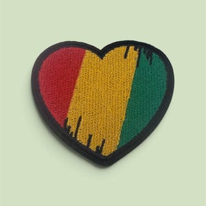 Op de afbeelding: Een hartvormige geborduurde patch met verticale strepen in rood, geel en groen. Het ontwerp heeft een zwarte rand en een verweerd verfdruppel-effect langs de onderkant van de gekleurde secties, wat zorgt voor een getextureerd uiterlijk.