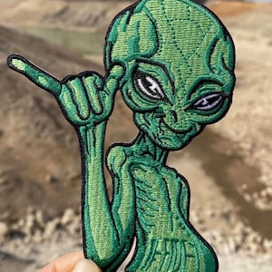 May include: An embroidered patch featuring a green alien character with a large head and dark eyes. The alien is making a shaka hand gesture with its right hand, with the thumb and pinky finger extended. The patch has a black border outline.