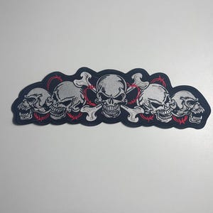 Op de afbeelding: Een geborduurde patch met een horizontale rij van vijf grijze schedels met gedetailleerde schaduwen. De middelste schedel is geplaatst over twee gekruiste witte botten. Dunne rode prikkeldraadaccenten zijn verweven tussen de schedels en botten.