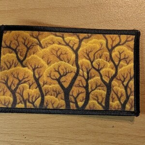 Puede incluir: Un parche rectangular con un diseño de bosque estilizado con árboles de color amarillo dorado y ramas negras. El parche está bordeado por un ribete negro. Una regla está al lado del parche.