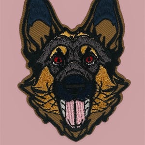 May include: Embroidered patch featuring a German Shepherd dog's head. The patch has a detailed design with brown, black, and gold colors. The dog's eyes are red, and its tongue is pink. The patch is set against a pink background.