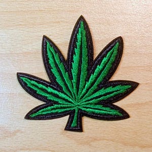 May include: Embroidered patch featuring a green cannabis leaf with black outlining. The leaf has detailed stitching and is set against a light wood grain background. A decorative item for clothing or accessories.