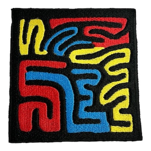 Può includere: Una toppa quadrata in tessuto nero con un design astratto colorato. Il design presenta linee intrecciate in rosso, giallo e blu. La toppa ha un aspetto strutturato, con le linee colorate che risaltano sullo sfondo nero.