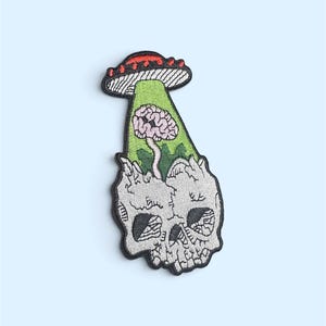 Op de afbeelding: Een geborduurde patch met een grijze schedel en een blootliggend brein aan de bovenkant. Een groene lichtstraal komt uit een rood-witte UFO boven de schedel en verlicht het brein. De patch heeft een zwarte rand en gedetailleerd stikwerk.