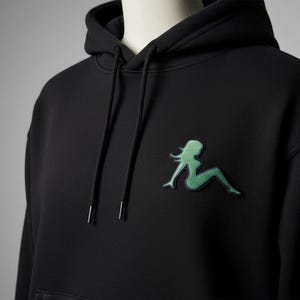 Op de afbeelding: Zwarte hoodie met een groen geborduurd grafisch ontwerp van een liggende vrouw op de borst. De hoodie heeft een capuchon met trekkoord en een kangoeroezak. De afbeelding is een silhouet met een getextureerde, gestikte uitstraling.