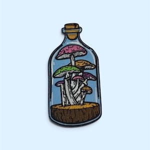 Op de afbeelding: Een geborduurde patch in de vorm van een glazen fles met een groep paddenstoelen erin. De paddenstoelen hebben roze, groene, gele en paarse hoeden met witte stelen, rustend op een bruine houten basis in de fles met een kurk.