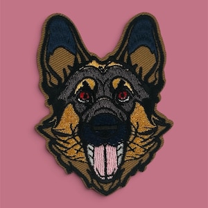 Op de afbeelding: Een geborduurde patch met de kop van een Duitse herder. Het ontwerp toont de karakteristieke bruine en zwarte vacht, puntige oren, donkere ogen en een roze tong die uit de bek hangt, alles omlijnd met donker garen.