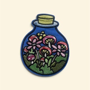 Op de afbeelding: Een ronde geborduurde patch in de vorm van een glazen fles met een gele kurk. De binnenkant toont een tafereel met roodhoedige paddenstoelen, roze bloemen en groen gebladerte tegen een blauwe achtergrond, omlijnd met donkerblauw garen.